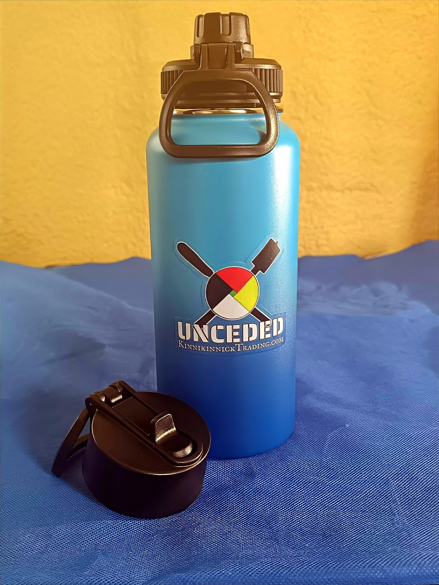 Water Is Life All Day Thermos Blue water bottle with UNCEDED logo and black lid on blue surface.